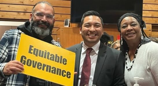 Edgar Franks (left) of Familias Unidas por la Justicia and Guillermo Rogel Jr. (center), Front and Centered's legislative advocate, smile and pose with a community member (right). Edgar holds a sign that reads "Equitable Governance."
