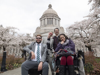 Members of BMAC mobility group standing and sitting in wheelchairs in front of capitol building in Olympia surrounded by cherry blossom trees. 